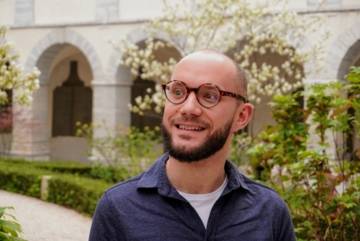 Portrait de Gianito, un jeune homme blanc et souriant. Il porte des lunettes rondes et une chemise bleu. Il regarde légèrement à gauche hors caméra.